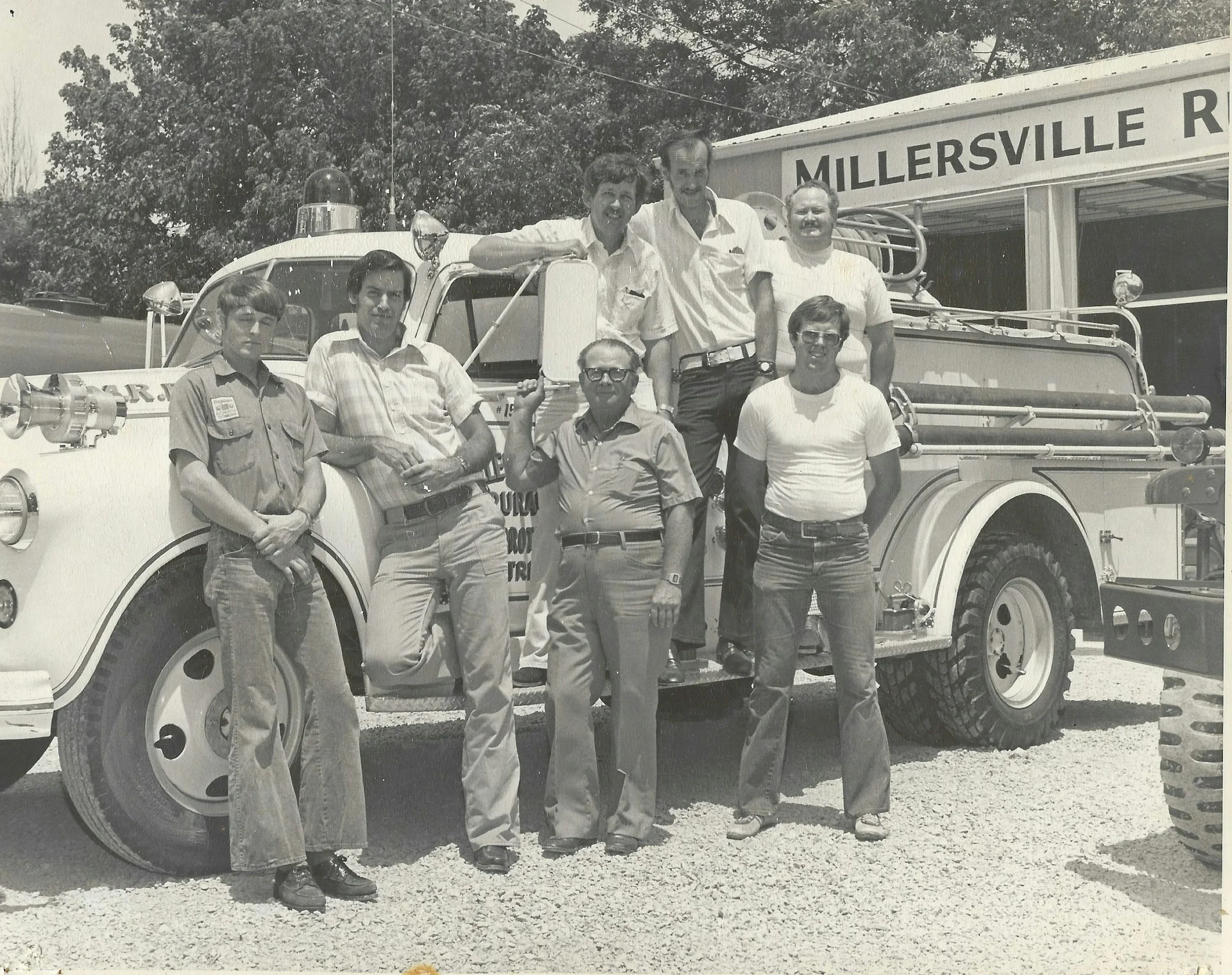 Historic black-and-white photo of seven adults posing beside a vintage fire engine in front of a building with “MILLERSVILLE R” visible on the sign.